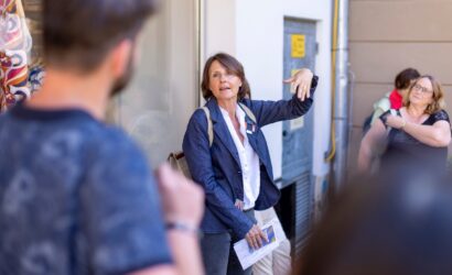 Eine Frau mit kurzer brauner Frisur erklärt gestikulierend etwas einer kleinen Gruppe auf der Straße. Sie trägt eine weiße Bluse, einen blauen Blazer und hat einen Rucksack auf dem Rücken. In der Hand hält sie Notizen und ein Magazin. Im Vordergrund ist ein Mann mit Bart unscharf zu sehen, im Hintergrund weitere Personen, darunter eine Frau mit schwarzem Shirt, die sich die Schulter reibt.