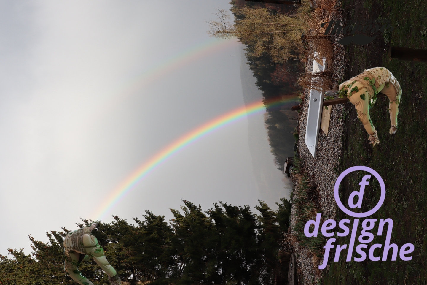 Landschaftsaufnahme mit doppeltem Regenbogen über einem bewaldeten Tal. Im Vordergrund befinden sich zwei humanoide Skulpturen, die mit Pflanzen und Stoff umhüllt sind. Rechts unten ist ein lilafarbenes Logo mit der Aufschrift „designfrische“ zu sehen.