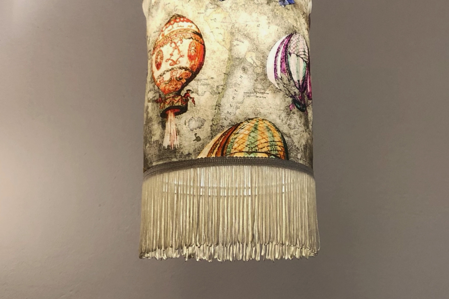 Eine dekorative Hängelampe mit nostalgischem Design, verziert mit Illustrationen bunter Heißluftballons auf einem Hintergrund in Vintage-Optik. Der Lampenschirm hat einen unteren Rand aus feinen, hängenden Fransen, die dem Objekt einen verspielten und eleganten Charakter verleihen.