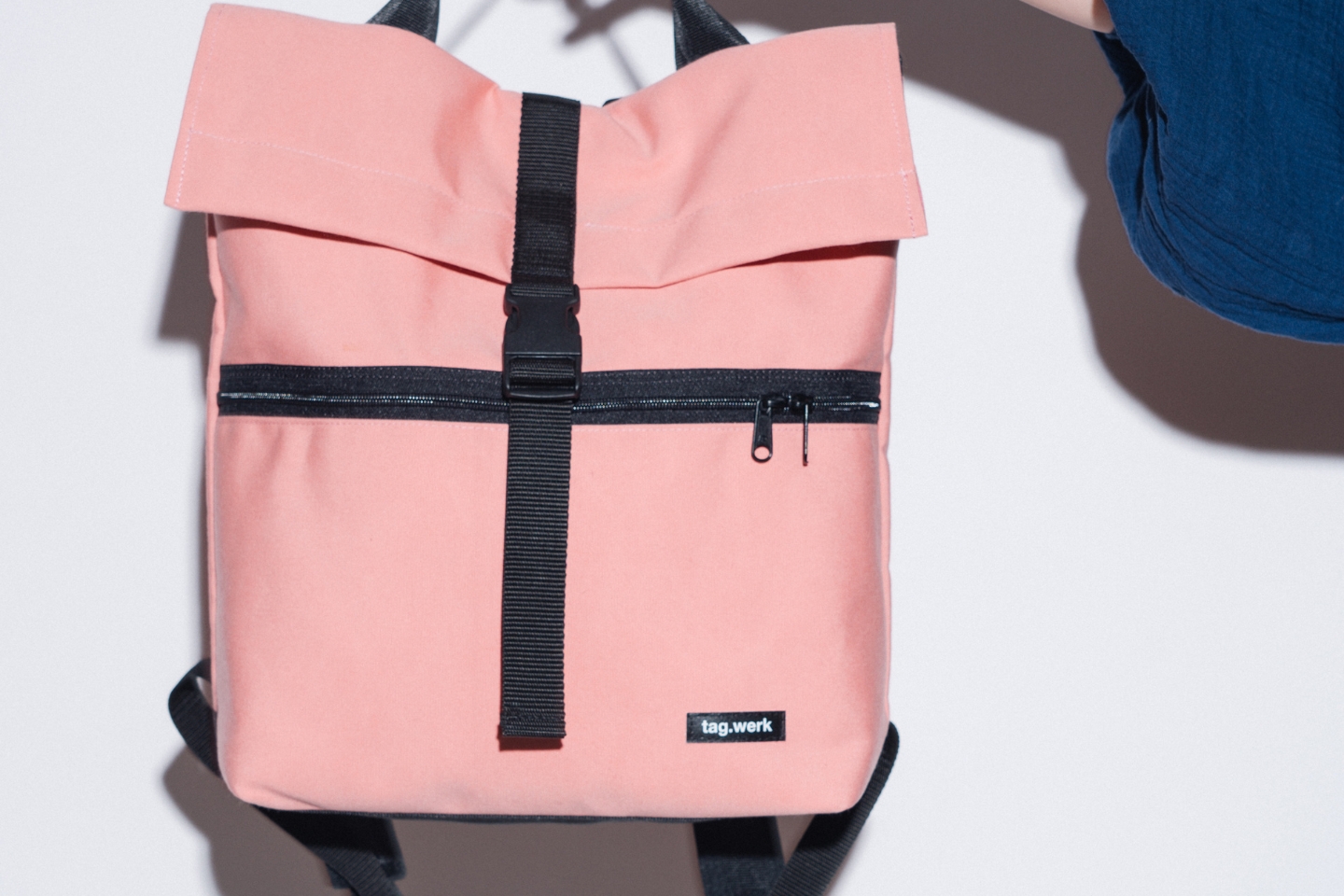 Rosa Rucksack mit schwarzem Reißverschluss, Schnalle und Trägern. Auf der Vorderseite befindet sich ein kleines Etikett mit der Aufschrift „tag.werk“. Eine Person in dunkelblauem Oberteil hält den Rucksack an einem Henkel hoch. Der Hintergrund ist hell mit starkem Schattenwurf.