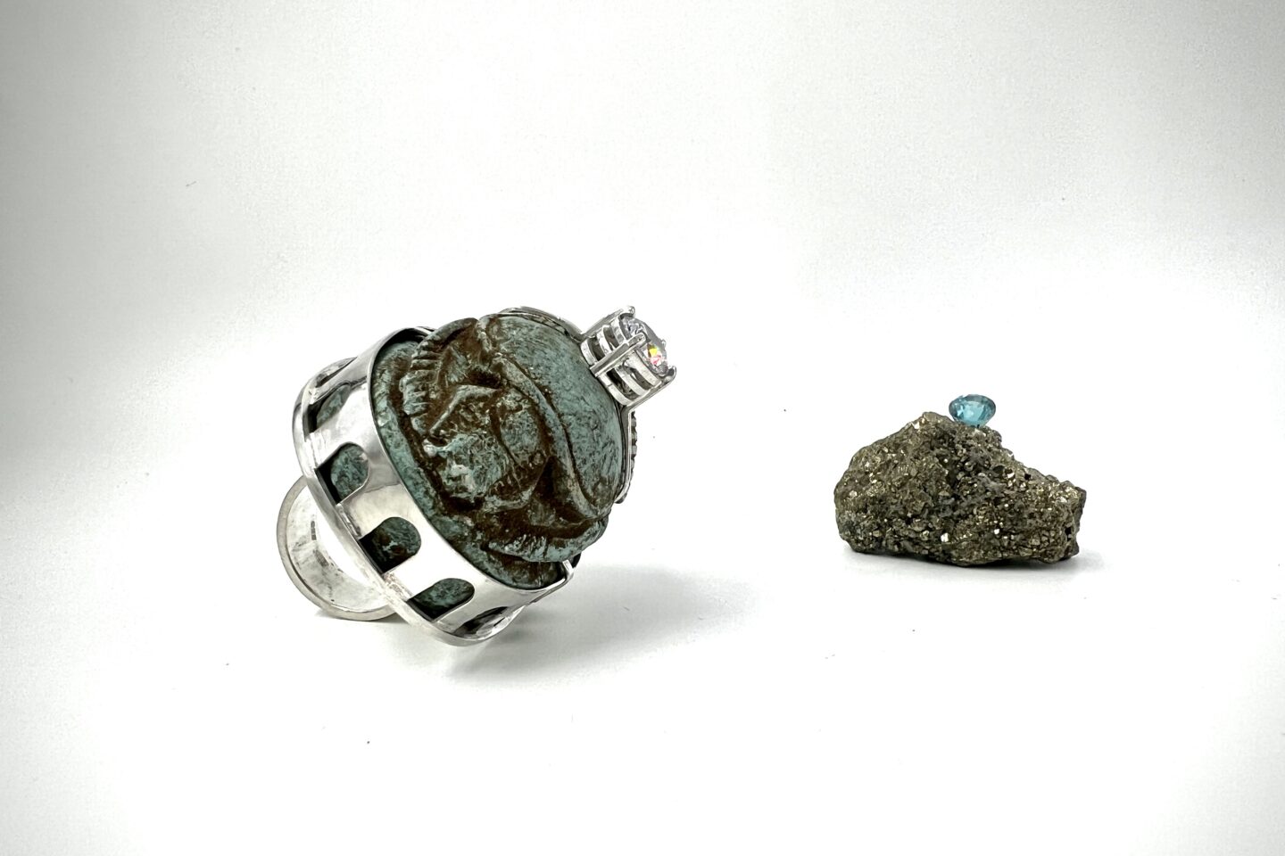 Zwei kunstvolle Schmuckstücke auf weißem Hintergrund – links ein Ring mit antik wirkendem Skarabäus-Stein in silberner Fassung, rechts ein glitzernder Rohstein mit einem eingefassten blauen Edelstein.