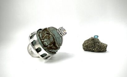 Zwei kunstvolle Schmuckstücke auf weißem Hintergrund – links ein Ring mit antik wirkendem Skarabäus-Stein in silberner Fassung, rechts ein glitzernder Rohstein mit einem eingefassten blauen Edelstein.