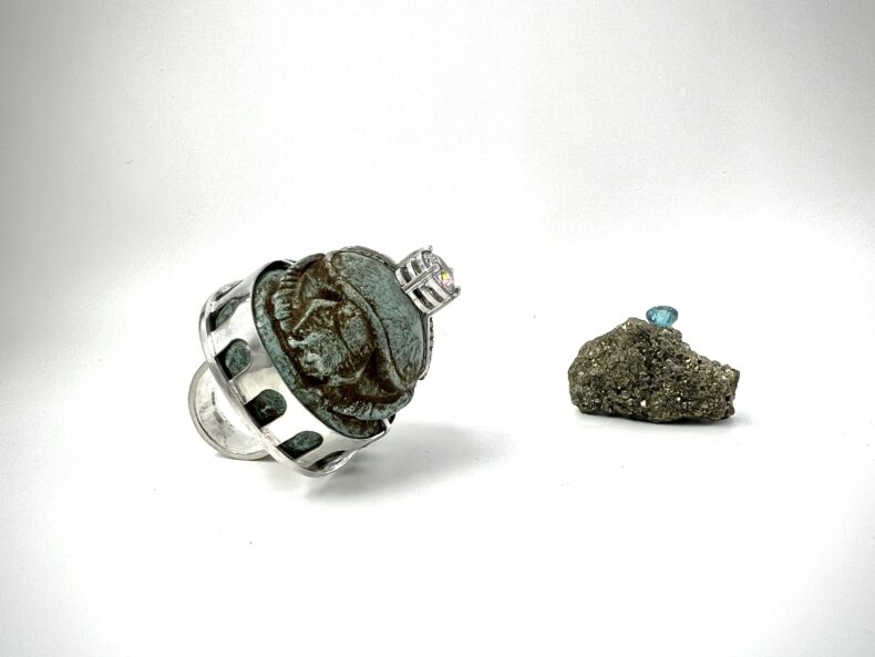 Zwei kunstvolle Schmuckstücke auf weißem Hintergrund – links ein Ring mit antik wirkendem Skarabäus-Stein in silberner Fassung, rechts ein glitzernder Rohstein mit einem eingefassten blauen Edelstein.