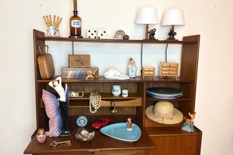 Vintage-Holzregal mit zahlreichen dekorativen Objekten, darunter eine Figur mit lila Flügelkleid, ein rotes Spielzeugauto, Schmuck, Porzellanfiguren, zwei Tischlampen, ein Strohhut, eine Matroschka-Puppe, Miniatur-Kommoden und ein alter Arzneiflasche mit der Aufschrift „Liniment B. Boirget“.