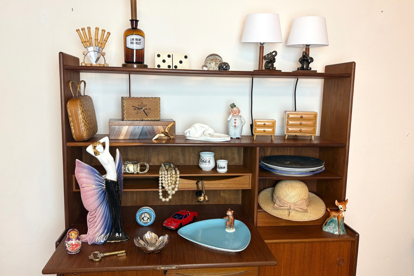 Vintage-Holzregal mit zahlreichen dekorativen Objekten, darunter eine Figur mit lila Flügelkleid, ein rotes Spielzeugauto, Schmuck, Porzellanfiguren, zwei Tischlampen, ein Strohhut, eine Matroschka-Puppe, Miniatur-Kommoden und ein alter Arzneiflasche mit der Aufschrift „Liniment B. Boirget“.