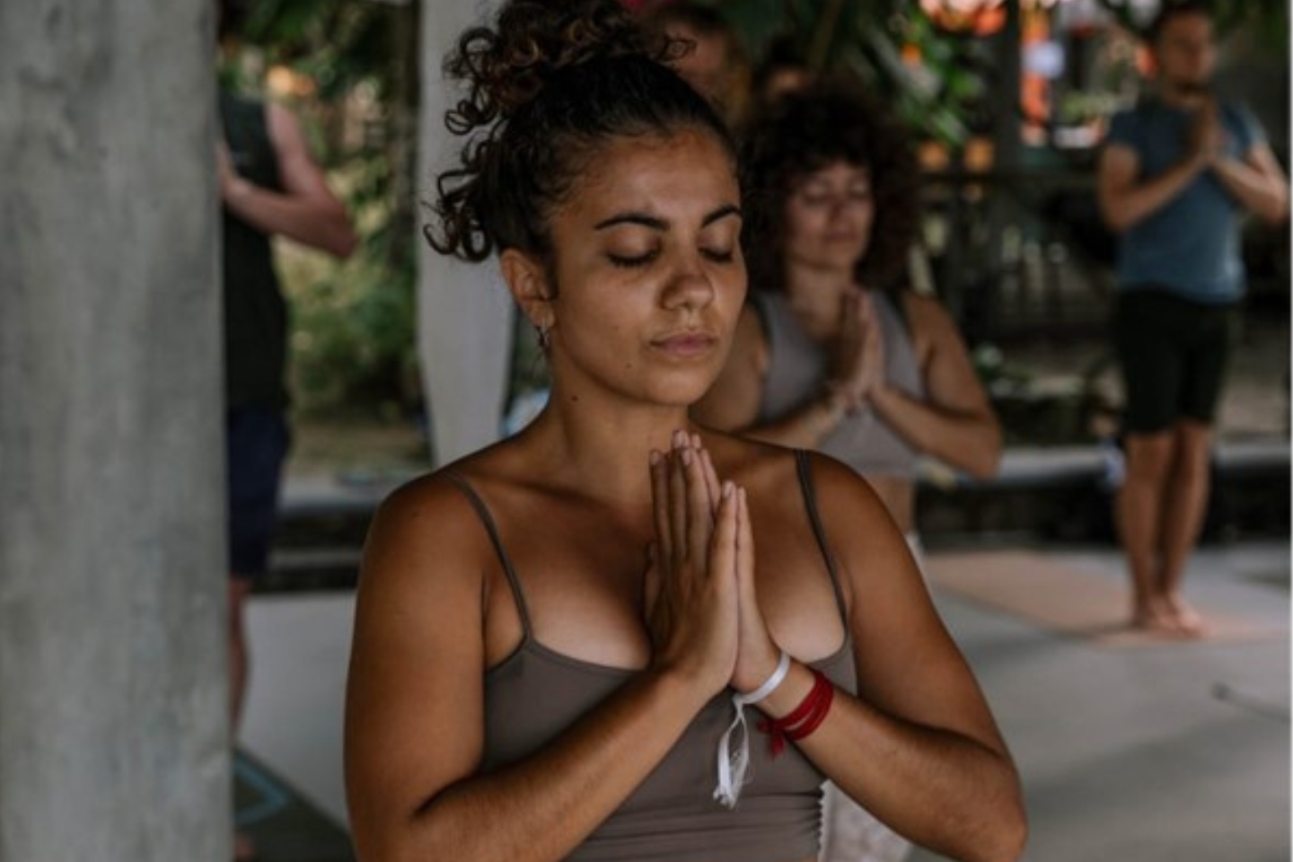 Eine junge Frau mit lockigem Dutt steht in Meditationshaltung mit geschlossenen Augen und gefalteten Händen in einem Yogastudio im Freien. Im Hintergrund sind weitere Personen in der gleichen Haltung zu sehen, umgeben von Pflanzen und Tageslicht.