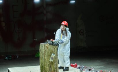 Eine Person in Schutzanzug, rotem Helm, Schutzbrille und Handschuhen zerschlägt mit einem Hammer ein elektronisches Gerät auf einem Baumstumpf in einem sogenannten „Rage Room“. Umherliegende Trümmerteile und grelles Licht verstärken die rohe, befreiende Atmosphäre.
