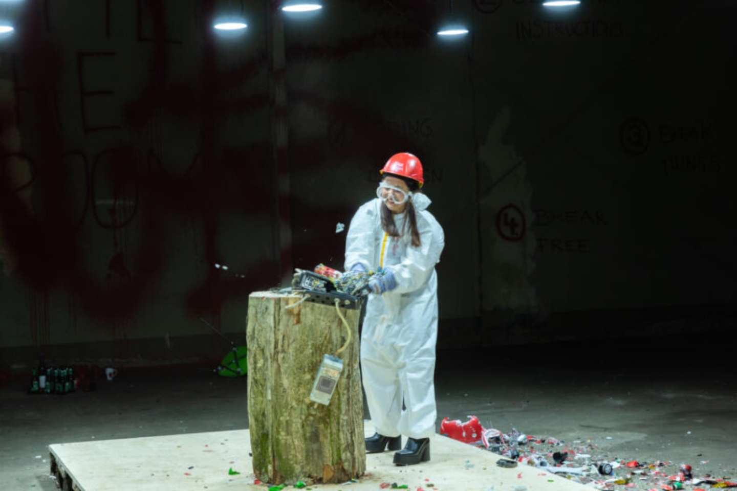 Eine Person in Schutzanzug, rotem Helm, Schutzbrille und Handschuhen zerschlägt mit einem Hammer ein elektronisches Gerät auf einem Baumstumpf in einem sogenannten „Rage Room“. Umherliegende Trümmerteile und grelles Licht verstärken die rohe, befreiende Atmosphäre.
