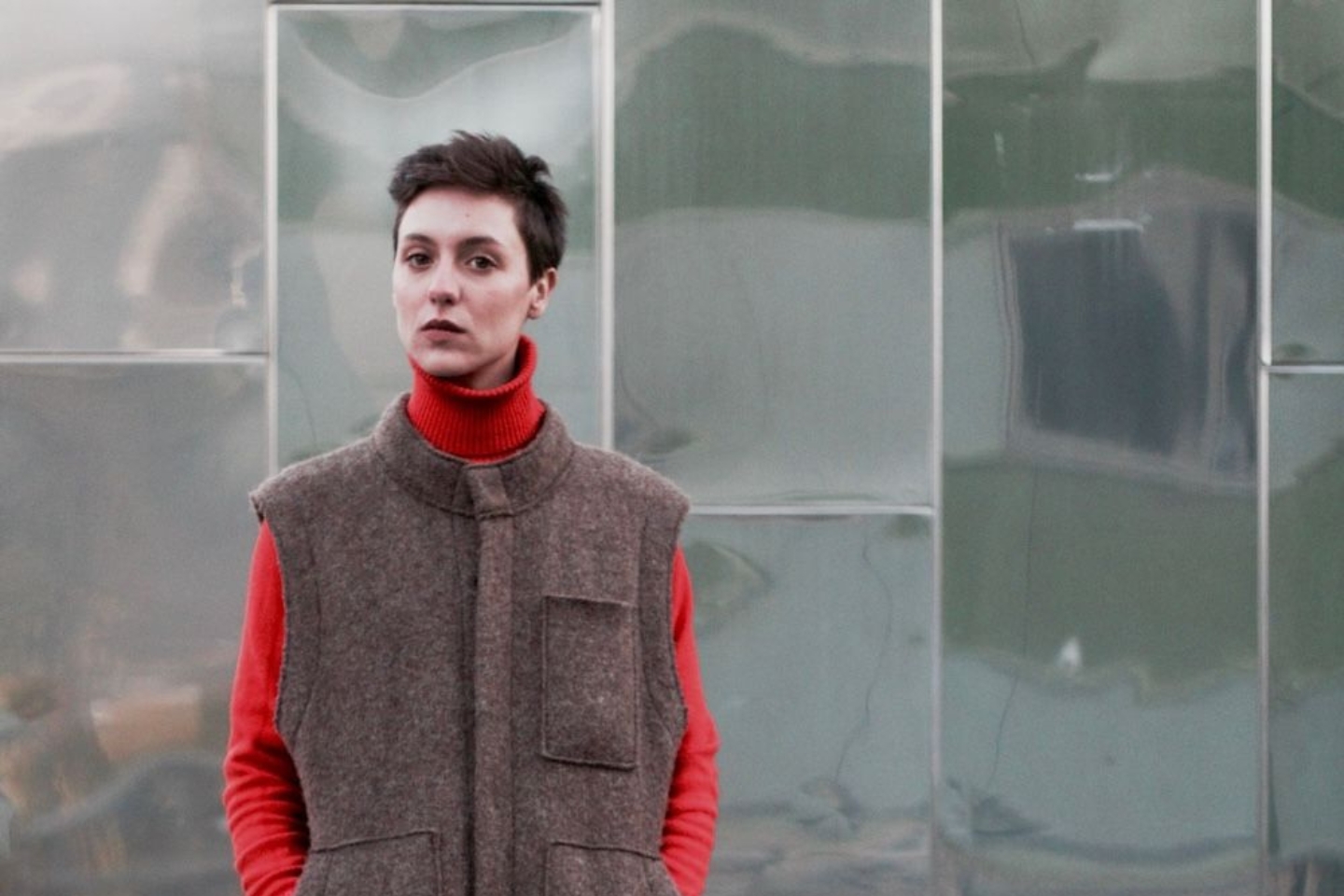 Eine Person mit kurzen dunklen Haaren steht vor einer metallisch glänzenden, leicht welligen Wand. Sie trägt einen roten Rollkragenpullover unter einer ärmellosen, braunen Wollweste mit aufgesetzten Taschen. Der Blick ist ruhig und direkt in die Kamera gerichtet.