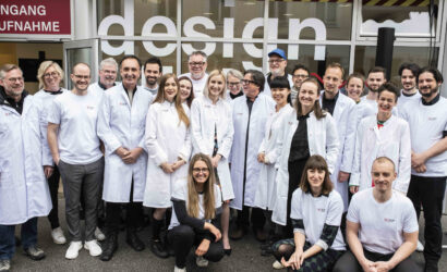 Eine Gruppe von Menschen in weißen Laborkitteln und T-Shirts mit Logo steht lächelnd vor einem Gebäude mit der Aufschrift „design“. Im Hintergrund ist ein Eingang mit einem Schild „Eingang Aufnahme“ zu sehen. Die Gruppe wirkt freundlich und einladend.